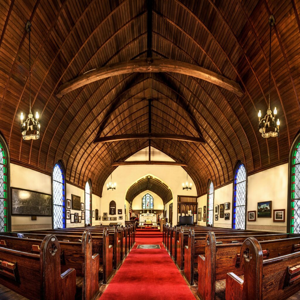 Inside view of a historic church featuring wooden arches, stained glass, and pew seating.